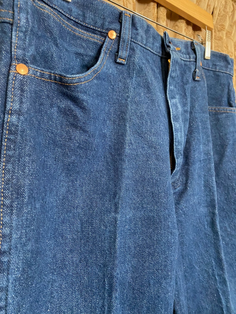 May include: Close-up of a pair of dark blue denim jeans. The jeans have copper-coloured rivets and stitching. The jeans are hanging on a wooden hanger.