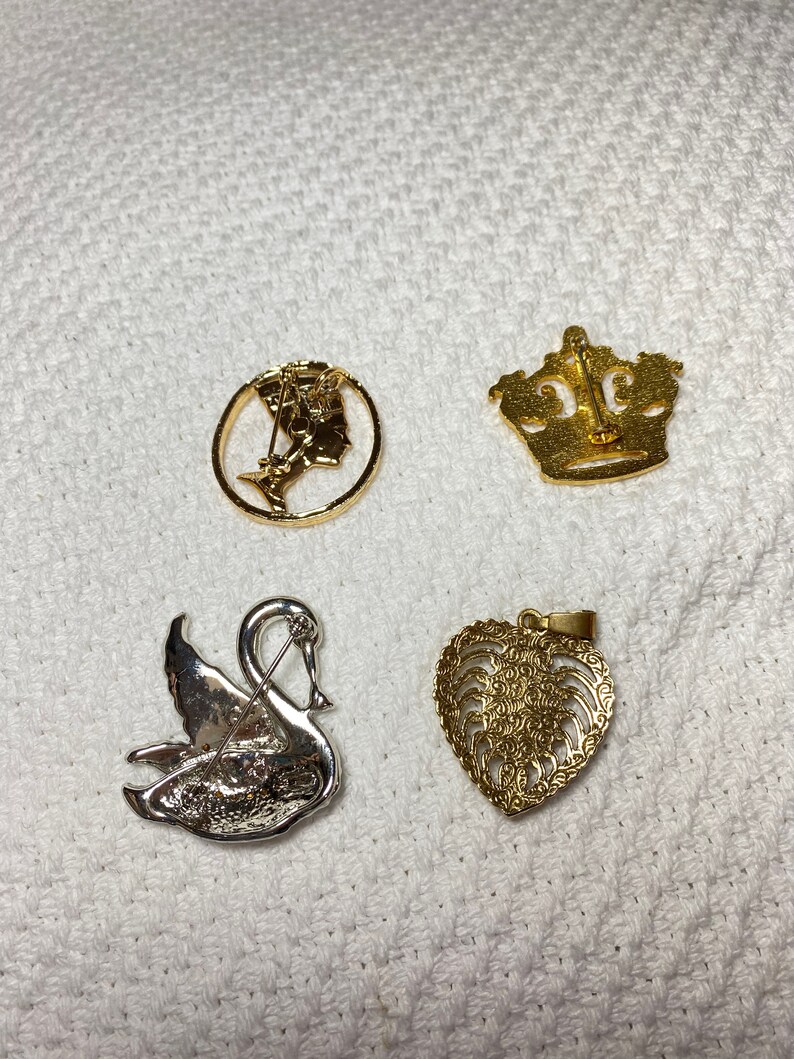 May include: Four vintage brooches and pendants. Includes a gold-tone crown pin, a gold-tone pendant with a heart-shaped filigree design, a silver swan pin, and a gold-tone pin with a profile of a woman.
