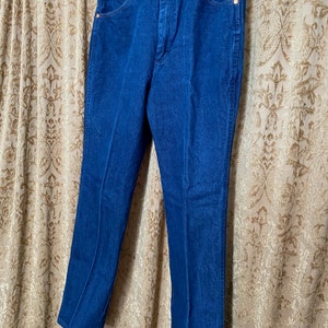 May include: A pair of blue denim jeans hanging on a wooden hanger. The jeans have a classic five-pocket design with copper-coloured rivets and a button closure. The denim appears to be a medium weight, with a slight fade.