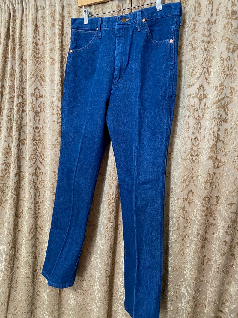 May include: A pair of blue denim jeans hanging on a wooden hanger. The jeans have a classic five-pocket design with copper-coloured rivets and a button closure. The denim appears to be a medium weight, with a slight fade.
