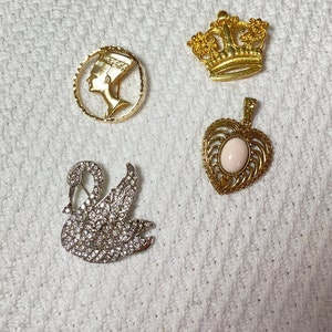 May include: A collection of vintage brooches and pendants. Includes a gold-tone Nefertiti profile brooch, a gold crown brooch, a silver swan brooch encrusted with rhinestones, and a gold heart pendant with a pink stone.