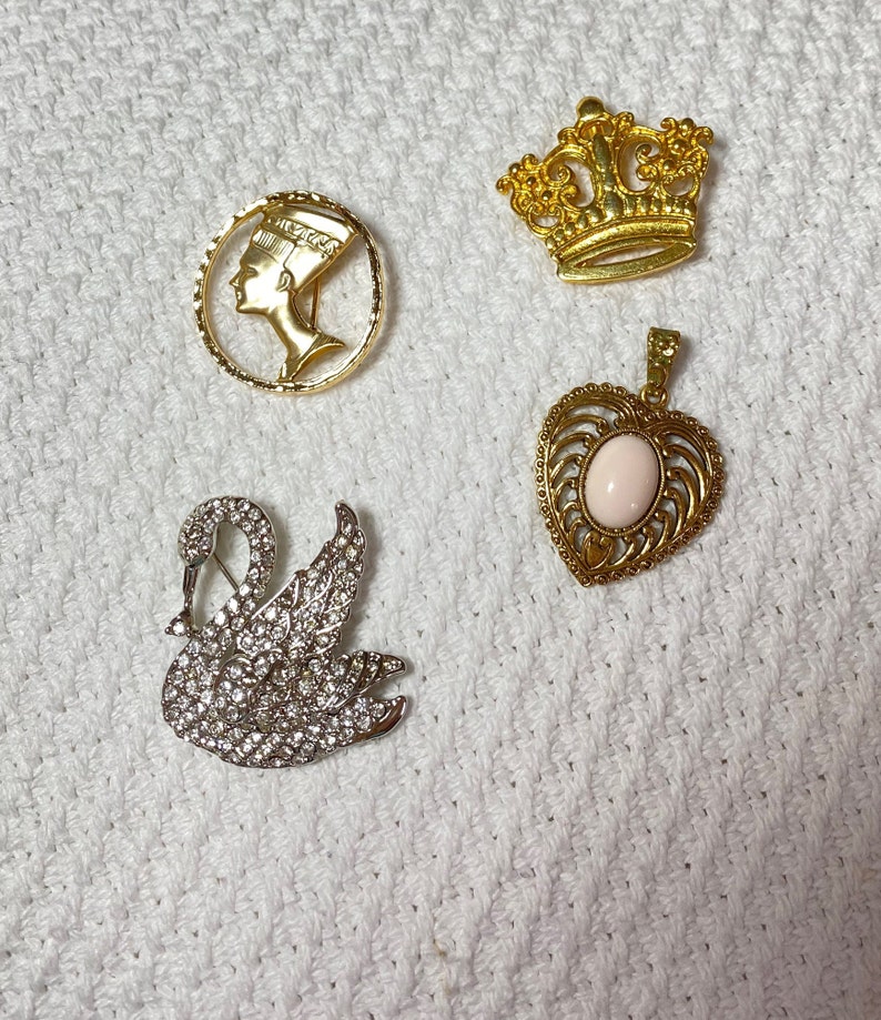 May include: A collection of vintage brooches and pendants. Includes a gold-tone Nefertiti profile brooch, a gold crown brooch, a silver swan brooch encrusted with rhinestones, and a gold heart pendant with a pink stone.
