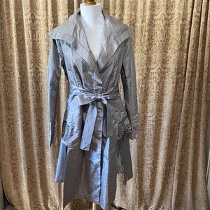 May include: A silver-grey trench coat with a wide collar and a matching tie belt. The coat has long sleeves and decorative patches on the lower front. It is displayed on a mannequin.