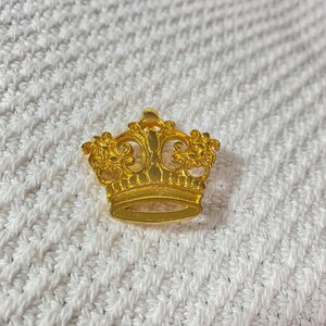 May include: A gold-colored crown pin with intricate filigree detailing. The crown features a classic design with a base and ornate top. The pin is set against a white, textured background.