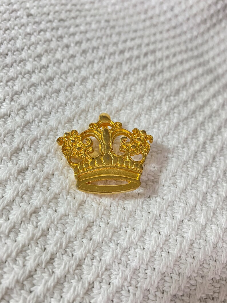 May include: A gold-colored crown pin with intricate filigree detailing. The crown features a classic design with a base and ornate top. The pin is set against a white, textured background.