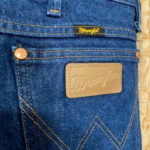 May include: Close-up of blue denim jeans with the Wrangler logo on a brown leather patch and a small tag. The jeans have copper rivets and gold stitching. The back pocket features a distinctive W-shaped design.