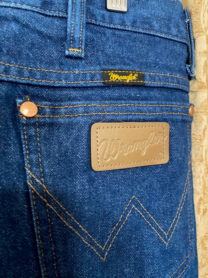 May include: Close-up of blue denim jeans with the Wrangler logo on a brown leather patch and a small tag. The jeans have copper rivets and gold stitching. The back pocket features a distinctive W-shaped design.