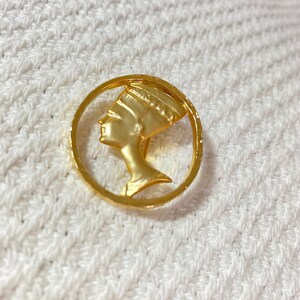 May include: A gold-colored brooch featuring a profile of a woman with an elaborate headdress, set within a circular frame. The brooch has a polished finish and is likely made of metal. The design is reminiscent of ancient Egyptian art.