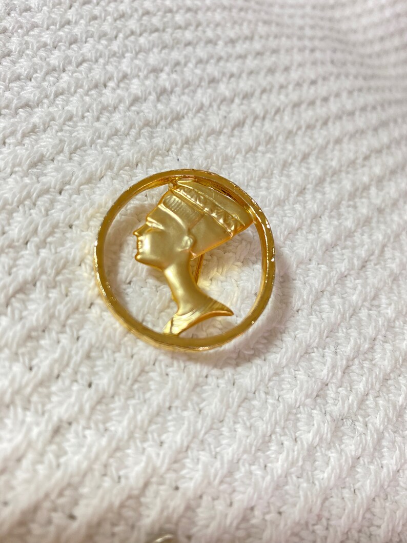 May include: A gold-colored brooch featuring a profile of a woman with an elaborate headdress, set within a circular frame. The brooch has a polished finish and is likely made of metal. The design is reminiscent of ancient Egyptian art.