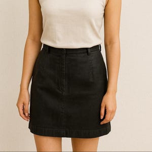 Op de afbeelding: Zwarte denim minirok met knoopsluiting en riemlussen. De rok is effen van kleur en heeft een klassieke, casual stijl. Gedragen met een crèmekleurige tanktop.