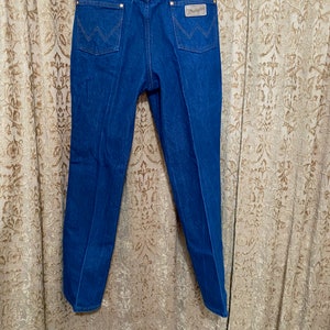 May include: Blue denim jeans with the Wrangler logo on the back pocket. The jeans are hanging on a wooden hanger against a patterned curtain. The jeans have a classic five-pocket design with copper-coloured rivets.