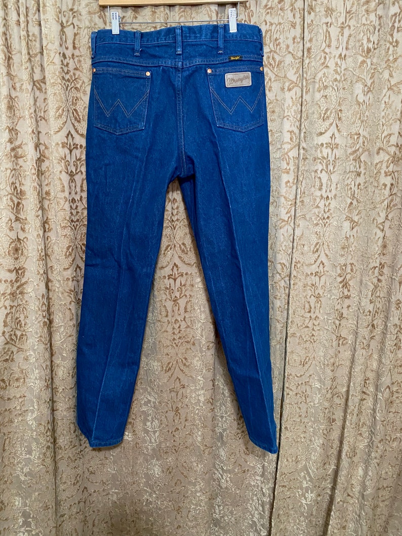 May include: Blue denim jeans with the Wrangler logo on the back pocket. The jeans are hanging on a wooden hanger against a patterned curtain. The jeans have a classic five-pocket design with copper-coloured rivets.