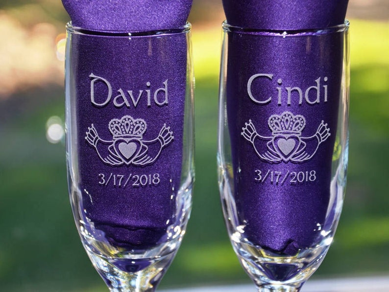 May include: Two clear glass champagne flutes containing deep purple fabric inserts. Each glass is etched with a Claddagh symbol, the name David or Cindi, and the date 3/17/2018. The design features two hands holding a heart topped with a crown.