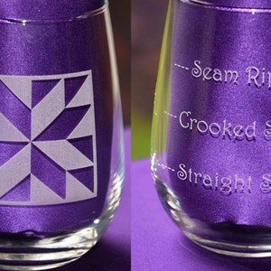 May include: A clear glass wine glass with a frosted design of a quilt block on the front. The back of the glass is etched with the words "Seam Ripper", "Crooked Stitch", and "Straight Stitch".