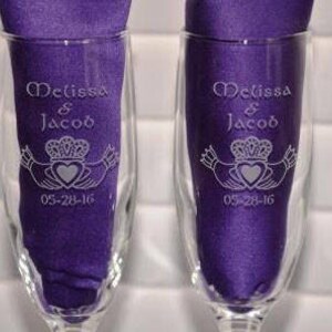 May include: Two clear glass champagne flutes, each containing a folded purple fabric napkin. The glass surface is etched with the names Melissa & Jacob, a Claddagh symbol featuring a heart and crown, and the date 05-28-16 in white lettering.