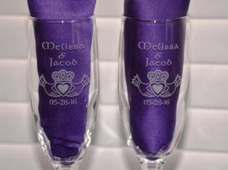 May include: Two clear glass champagne flutes, each containing a folded purple fabric napkin. The glass surface is etched with the names Melissa & Jacob, a Claddagh symbol featuring a heart and crown, and the date 05-28-16 in white lettering.