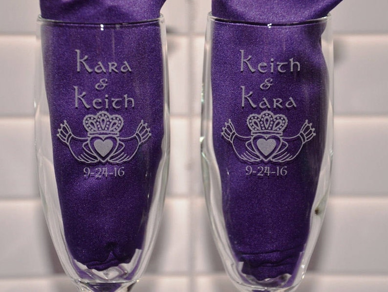 May include: Two clear glass champagne flutes featuring etched designs. One glass is engraved with the names Kara & Keith, the other with Keith & Kara, both including a Claddagh symbol and the date 9-24-16. Each glass contains a purple fabric insert.