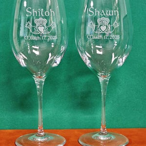 May include: Two clear glass wine glasses featuring etched designs. Each glass displays a Claddagh symbol, the name Shiloh or Shawn, and the date March 17, 2025. The glasses have a long stem and a wide circular base, standing upright on a flat surface.