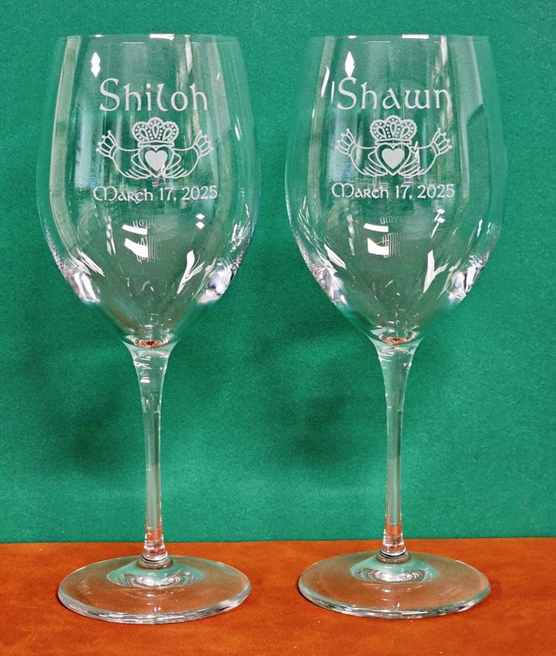 May include: Two clear glass wine glasses featuring etched designs. Each glass displays a Claddagh symbol, the name Shiloh or Shawn, and the date March 17, 2025. The glasses have a long stem and a wide circular base, standing upright on a flat surface.