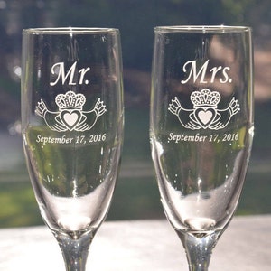 May include: Two clear glass champagne flutes featuring white etched designs. One glass is engraved with Mr., a Claddagh symbol, and September 17, 2016. The other glass is engraved with Mrs., a Claddagh symbol, and September 17, 2016.
