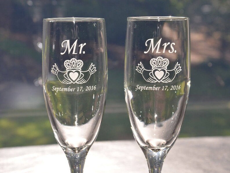 May include: Two clear glass champagne flutes featuring white etched designs. One glass is engraved with Mr., a Claddagh symbol, and September 17, 2016. The other glass is engraved with Mrs., a Claddagh symbol, and September 17, 2016.