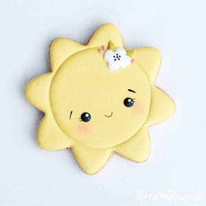 May include: A yellow sun-shaped cookie with a smiling face, rosy cheeks, and a small white flower with pink accents. The cookie has a detailed design and is likely a decorative treat.