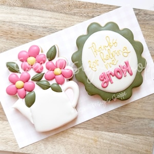May include: Two decorated sugar cookies. One cookie is green with a white circle and the text "Thanks for helping me grow". The other cookie is white and shaped like a watering can with pink flowers and green leaves.