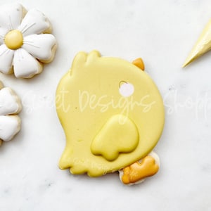 May include: A yellow chick cookie decorated with white icing and a pink eye. The cookie is on a white surface.