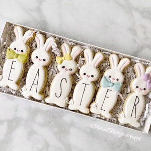 Puede incluir: Un conjunto de seis galletas de azúcar de conejito blanco decoradas con colores pastel y la palabra "EASTER" deletreada. Las galletas están dispuestas en una caja blanca con papel triturado.