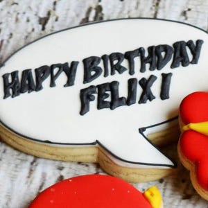 May include: A decorated cookie in the shape of a speech bubble with the words "HAPPY BIRTHDAY FELIX!" in black icing. Additional cookies are visible, including a red and yellow design. The cookies are on a light-colored surface.