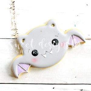 May include: A gray and purple cookie decorated to look like a cartoon bat with big eyes and fangs.