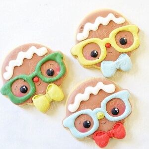 May include: Three gingerbread cookies decorated with icing. Each cookie has a different colored bow tie and glasses. The glasses are green, yellow, and blue. The cookies are on a white background.