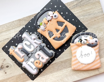 May include: Three Halloween-themed cookies on a wooden board. One cookie is a jack-o'-lantern, another says "Trick or Treat", and the third is a candy bag with the word "boo!". The cookies are decorated with orange, black, white, and gray icing.
