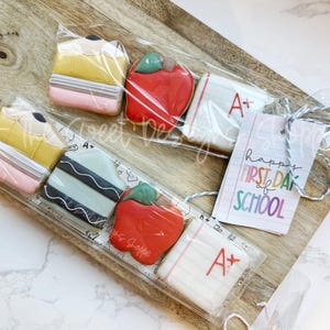 May include: A set of six decorated sugar cookies with a back to school theme. The cookies are decorated with images of pencils, apples, and a notebook with an A+ grade. The cookies are packaged in clear cellophane bags with a tag that reads "Happy First Day of School".