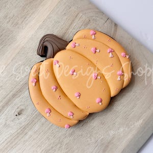 May include: A pumpkin-shaped cookie with a light orange base, decorated with small pink mushroom accents. The cookie has a dark brown stem and is placed on a wooden surface. The Sweet Designs Shoppe is written on the surface.