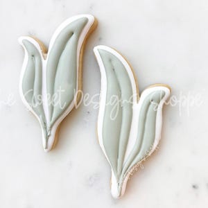 May include: Two leaf-shaped cookies with light green icing and white accents. The cookies have a light brown base and are decorated with a detailed design. The cookies are on a white marble surface.