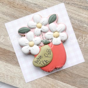 May include: A pink and white sugar cookie decorated with a bouquet of flowers in a red pot. The pot has the text "Mrs. Tea Pass" written on it.