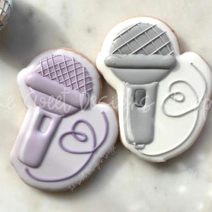 May include: Two decorated sugar cookies shaped like microphones. One cookie is white with a gray microphone and the other is light purple with a purple microphone. Both cookies have a white icing swirl.