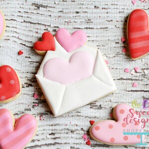 May include: A set of Valentine's Day sugar cookies decorated with pink and red icing. The cookies are shaped like hearts and envelopes. One cookie has the text "the sweet designs SHOPPE LLC" on it.