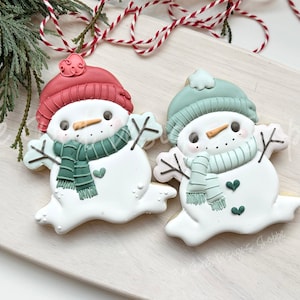 May include: Two snowman-shaped cookies with white frosting, each wearing a hat and scarf. One has a red hat and green scarf, the other a green hat and scarf. Both have a carrot nose and black eyes. Christmas-themed.
