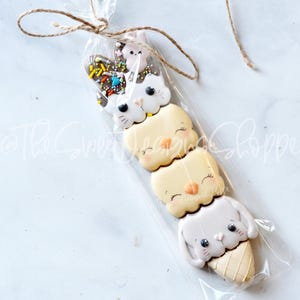 May include: A set of four decorated sugar cookies in a clear plastic bag tied with twine. The cookies are shaped like ice cream cones and feature animal faces. The top cookie is a white cat, the next is a yellow chick, the next is a yellow chick, and the bottom is a white bunny.