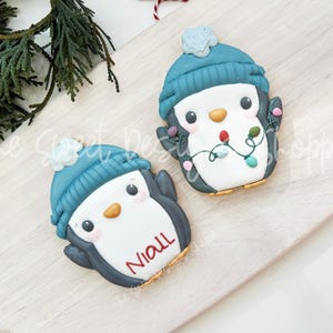 May include: Two penguin-shaped cookies with blue hats and festive lights. One cookie has the name "Niall" written on it. The cookies are decorated with white, gray, and orange icing, and are on a light-colored wooden surface.