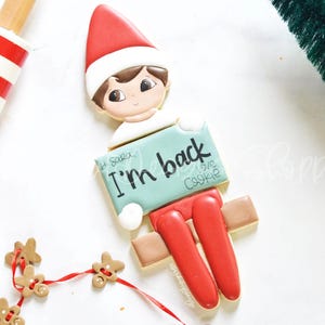 May include: A gingerbread elf cookie decorated with red, white, and brown icing. The elf is holding a sign that says "I'm back I love cookie".