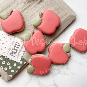 May include: Pink apple-shaped cookies with green stems and leaves are arranged on a wooden cutting board. A tag with the text "I couldn't have PICKED a better teacher" is also present. The cookies are likely for a teacher appreciation gift.