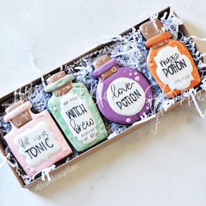 May include: A box of four potion-shaped cookies with various colors and labels. The cookies are pink, green, purple, and orange, with text that reads "bat wing TONIC", "Witch Brew", "love POTION", and "magic POTION". The cookies are in a brown box with white paper filler.