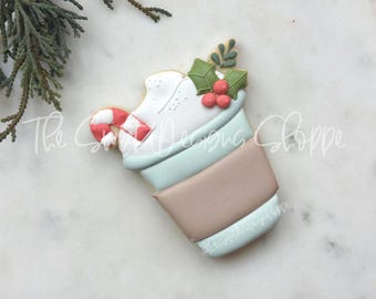 Puede incluir: Una galleta de azúcar decorada con forma de taza de chocolate caliente con crema batida, un bastón de caramelo y una rama de acebo con bayas rojas.