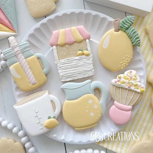 May include: A set of six decorated sugar cookies with a lemonade theme. The cookies are decorated with yellow, pink, white, and blue icing. The cookies include a pitcher, a glass, a lemon, a lemonade stand, and a mason jar.