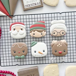 May include: Christmas-themed cookies arranged on a cooling rack. The cookies feature Santa Claus, an elf, Mrs. Claus, a gingerbread person, a snowman, and a reindeer. The cookies are decorated with various colors and details.