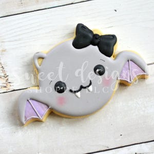 May include: A gray bat cookie with purple wings and a black bow. The bat has a cute face with black eyes and white teeth.