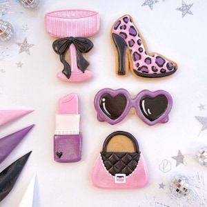May include: An assortment of decorated cookies in the shape of fashion accessories. The cookies include a pink lamp with a black bow, a high heel with a leopard print, heart-shaped sunglasses, a lipstick, and a purse.